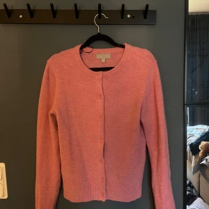 Rosa stickad kofta från H&M - Mysig rosa stickad kofta från H&M i storlek XS. Koftan har rund hals, långa ärmar och knäppning med matchande rosa knappar framtill. Perfekt att slänga på sig när det är lite kyligt ute. Ribbstickade muddar vid ärmslut och nederkant.
