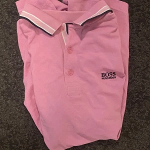 Rosa Hugo Boss pikétröja M - Snygg rosa pikétröja från Hugo Boss med klassisk krage och knäppning framtill. Regular fit och broderad logga på bröstet. Tröjan har korta ärmar och är tillverkad i mjuk bomull som andas. Perfekt för dig som vill ha en clean och stilren look.