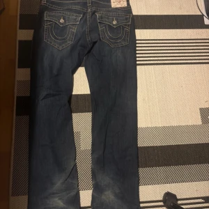 True Religion  jeans blå, straight/baggy - Säljer ett par True Religion Ricky jeans i blå tvätt med kontrastsömmar och snygga fickdetaljer bak. Modellen är relaxed straight med klassisk femficksdesign och markerade ficklock. Perfekt för dig som gillar street och premium denim.