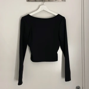 Svart långärmad topp med spets - Snygg svart långärmad topp med croppad passform och bred urringning. Baksidan har ett transparent spetslager med blommigt mönster som ger en extra detalj. Perfekt att styla för en trendig look. (Aldrig använd)