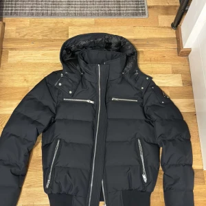 Svart puffer bomberjacka Moose Knuckles - Snygg svart puffer bomberjacka från Moose Knuckles med hög krage, avtagbar huva och flera silvriga dragkedjor. Jackan har ribbade muddar och logga på ärmen. Perfekt för kalla dagar när du vill hålla stilen.