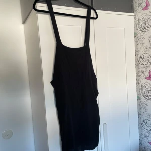 Svart kort hängselklänning med dragkedja - Svart kort klänning med breda axelband och en cool ficka med dragkedja på sidan. Klänningen har en enkel, loose passform och är tillverkad i mjuk bomull.  Klänningen har gått upp i några sömmar men lätt att laga, där av billigt pris. Storlek 42 men passar 38 bäst.