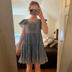 Blå blommig kortklänning med volang - Supersöt blå kortklänning med vita blommor och volangärmar. Klänningen har fyrkantig ringning, dekorativa broderier och ett flowy fall. Perfekt för varma dagar och har en riktigt bohemisk vibe.