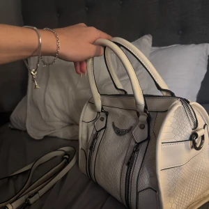 Vit handväska  - Snygg vit handväska med ormmönstrad struktur och svarta detaljer. Väskan har dubbla handtag, avtagbar axelrem och två dragkedjor framtill. Dekorativ metallvinge på framsidan ger extra edge. Perfekt storlek för allt du behöver till vardags.