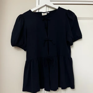 Mörkblå blus med knyt från Gina Tricot - Supersöt svart blus från Gina Tricot med puffiga korta ärmar och tre knyt framtill. Blusen har en lös passform och volang nedtill som ger en chill och trendig vibe. Perfekt att styla med jeans eller kjol för en snygg look.