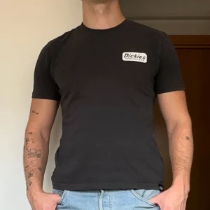 Svart Dickies t-shirt - Svart t-shirt från Dickies med vit logga på bröstet och flera coola tryck i vitt på ryggen. Klassisk passform och rund hals. Perfekt för dig som gillar streetwear och vill ha en enkel men snygg look. Gjord i mjuk bomull.