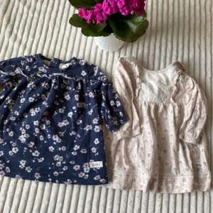 2 blommiga klänningar från Newbie, barn 68 - Två söta kortklänningar från Newbie. En marinblå med ljusrosa och vita blommor, volangdetalj och lång ärm. Den andra är ljusbeige med små rosa blommor, spets på bröstet och volang vid axlarna. Båda har mjukt bomullstyg och är perfekta för vår och sommar. Storlek 68.