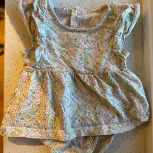 Blommig babyklänning från Carter's, barn 68 - Supersöt babyklänning från Carter's i bomull med blommigt mönster i pastellfärger som rosa, gul och grön. Klänningen har volangdetaljer vid axlarna och praktiska tryckknappar i grenen för enkel påklädning.