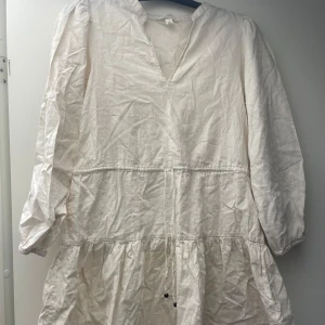 Vit långärmad klänning i linne - Luftig vit klänning i linne med långa ärmar och v-ringning. Klänningen har snörning i midjan och volangdetaljer nedtill för en avslappnad och trendig look. Perfekt för varma dagar och enkel att styla med accessoarer.