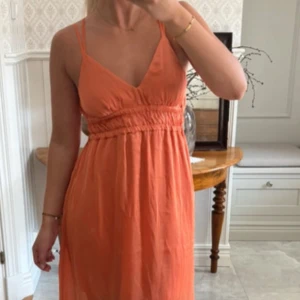 Orange sommarklänning med v-ringning - Snygg orange klänning med smala axelband och v-ringning. Modellen är luftig och har rynkade detaljer under bysten för en skön passform. Perfekt för varma dagar och ger en fräsch look. Materialet känns lätt och svalt mot huden.
