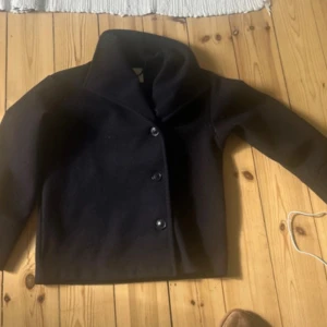 Svart kappa  från Gina Tricot - Stilren svart rock från Gina Tricot i ullblandning. Jackan har en rak, boxig passform, bred krage och knäpps med tre knappar framtill. Perfekt för dig som gillar en klassisk look med modern twist.