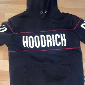 Svart HOODRICH hoodie med röd detalj - Svart hoodie från HOODRICH med stor vit logga över bröstet och röda linjer som ger en sportig vibe. Tröjan har huva med snörning och en stor magficka. Snygga vita tryck på ärmarna för extra streetstyle.