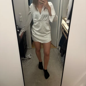 Vit omlottklänning från Zara - Snygg vit kort omlottklänning från Zara i glansigt satintyg. Klänningen har långa ärmar, v-ringning och markerad midja som ger en riktigt fräsch och trendig look. Perfekt för dig som gillar stilrena och eleganta outfits.