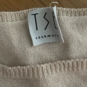 Beige cashmere linnetopp från TSE - Superskön och lyxig beige linnetopp från TSE i 100% kashmir. Enkel och stilren design med rund halsringning och ribbade kanter. Perfekt att styla med jeans eller kjol för en clean look.