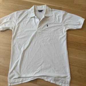 Vit pikétröja från Polo Ralph Lauren - Klassisk vit pikétröja från Polo Ralph Lauren med broderad logga på bröstet. Tröjan har krage, knäppning med två knappar och är tillverkad i 100% bomull. Perfekt för en clean och stilren look.