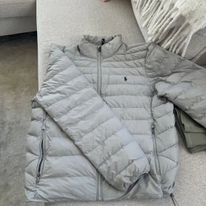 Grå dunjacka från Polo Ralph Lauren - Snygg grå dunjacka från Polo Ralph Lauren med klassisk quiltad design och diskret logga på bröstet. Jackan har dragkedja framtill, två sidofickor med dragkedja och hög krage. Perfekt för kyliga dagar och enkel att matcha med allt.