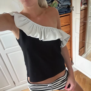 Svart one shoulder topp med volang - Snygg svart topp med en axel och stor vit volang som går över axeln och ryggen. Toppen är ärmlös och har en oneshoulder-design som ger en cool och trendig look. Perfekt för dig som vill sticka ut lite extra. Storlek L men skulle säga att den är en M/s