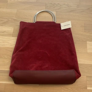 Vinröd axelväska från Carin Wester - Säljer en snygg vinröd axelväska från Carin Wester i mocka och skinn. Väskan har silvriga metallhandtag, avtagbar axelrem och en matchande innerpåse. Perfekt för dig som gillar stilrena accessoarer med en twist. Väskan är i nyskick med prislapp och nypris är 499kr. Pris går att diskutera!🍷👜