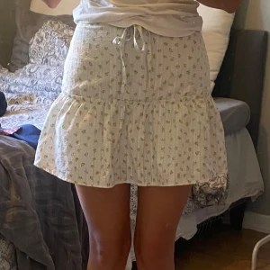 Vit blommig volangkjol med snörning - Supersöt kort kjol i vitt med små blommor och volang nertill. Kjolen har snörning i midjan och är gjord i ett lätt bomullstyg, perfekt för varma dagar. 