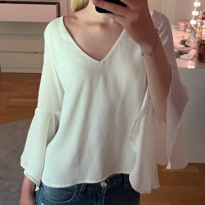 Vit blus med öppen rygg från Zara - Säljer en vit blus från Zara med v-ringning och vida, fladdriga ärmar. Baksidan har en snygg öppen rygg-detalj som gör toppen extra speciell. Perfekt att styla med jeans eller kjol för en trendig look.