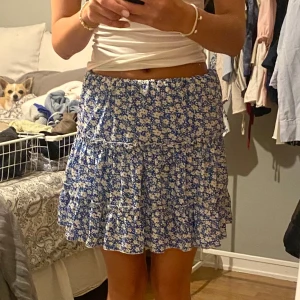 Blommig blå volangkjol - Supersöt kort kjol med blå botten och vita blommor. Kjolen har volanger och resår i midjan för en bekväm passform. 