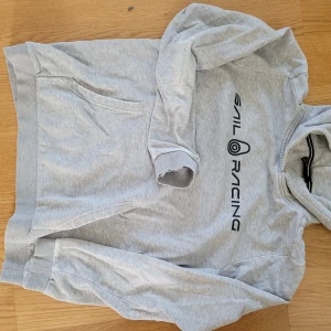 Grå hoodie från Sail Racing - Ljusgrå hoodie från Sail Racing med svart logga på bröstet. Klassisk känguruficka framtill, huva och långa ärmar. Mjuk insida och ribbade muddar som är slitna, se bild. . Perfekt för chill dagar eller när du vill ha en enkel och stilren look.