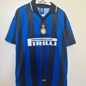 Inter Milan retro fotbollströja L - Klassisk Inter Milan fotbollströja i blått och svart med vertikala ränder, broderat klubbmärke och gul stjärna på bröstet. Tröjan har svart krage med vit detalj, V-ringning och stor Pirelli-logga framtill. Tillverkad i lätt och ventilerande material.