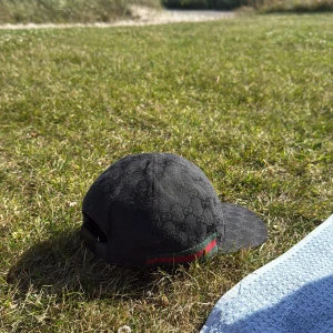 Svart Gucci keps med GG-mönster - Snygg svart keps från Gucci med diskret GG-monogram över hela kepsen och klassiska gröna och röda ränder på sidan. Kepsen har böjd skärm och justerbar baksida. Perfekt för dig som vill ha en lyxig och stilren accessoar.