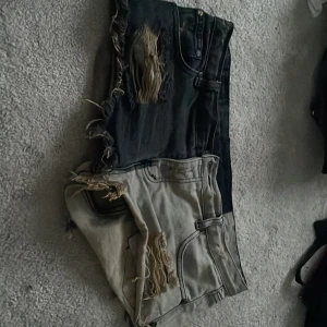 Tvåfärgade one teaspoon jeansshorts  - Unika jeansshorts från one teaspoon  med tvåfärgad design – ena sidan svart, andra beige. Råa slitningar och fransiga kanter ger en edgy vibe. Klassisk femficksmodell med låg midja och knappgylf. Perfekta för dig som vill sticka ut i sommar.