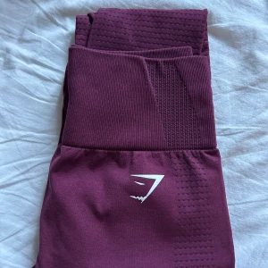 Vinröda seamless leggings från Gymshark - Säljer ett par vinröda seamless leggings från Gymshark med hög midja. De är tillverkade i ett stretchigt syntetmaterial som sitter tight och är perfekta för träning. Gymshark-logga under midjan och på vaden. Använda endast ett fåtal gånger. Hör gärna av er vid minsta lilla fråga! Säljs även som paket med dem andra träningsbyxorna. Hör av er till mig om ni är intresserade av paketspris. 
