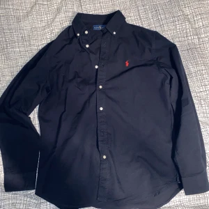 Mörkblå skjorta från Ralph Lauren M - Klassisk mörkblå skjorta från Ralph Lauren med vita knappar och röd broderad logga. Storleken är M men passar även närliggande storlekar. Endast testad men aldrig använd utanför hemmet. Vid frågor tveka inte om att höra av!