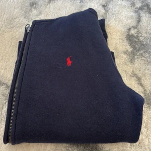 Marinblå hoodie från Polo Ralph Lauren - Snygg marinblå hoodie från Polo Ralph Lauren med dragkedja och huva. Klassisk röd logga broderad på bröstet och praktiska fickor framtill. Tillverkad i mjuk bomullsmix, perfekt för chill dagar eller när du vill ha en clean streetstyle.