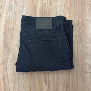 Svarta stretchjeans från Hugo Boss - Snygga svarta jeans från Hugo Boss med regular fit och stretch för extra komfort. Klassisk femficksmodell med diskret Boss-logga på bakfickan och läderpatch i midjan. Perfekta till en stilren look och passar till det mesta.
