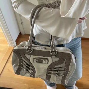 Guess väska - Snygg handväska från Guess i grått och beige med reptilstruktur. Väskan har dubbla handtag, dekorativa spännen och metallplatta framtill. Perfekt storlek för att få plats med allt du behöver och ger en cool touch till din outfit.