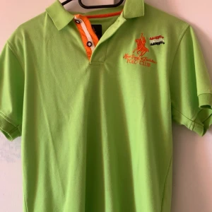 Grön pikétröja från Polo Club - Snygg grön pikétröja från Polo Club med orange detaljer och broderad logga på bröstet. Klassisk krage, korta ärmar och knappar framtill. Perfekt för en sportig och fräsch look. Materialet känns mjukt och bekvämt, troligen bomull.