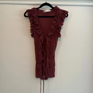 Zara topp  - Supersöt vinröd ribbad topp från Zara med volangdetaljer längs axlar och urringning. Toppen är ärmlös och har knytband framtill. 