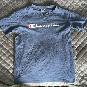 Blå Champion t-shirt i bomull XL - Snygg blå t-shirt från Champion i storlek XL. Klassisk modell med korta ärmar och rund hals. Stor logga i vitt och rött framtill. Tillverkad i mjuk bomull som är skön att ha på sig. Perfekt för en avslappnad streetstyle-look.