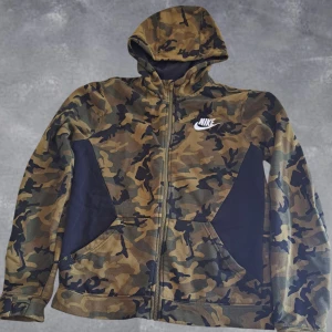 Nike camo zip hoodie - Nike zip hoodie med camouflagemönster i gröna och bruna toner. Jackan har huva, hel dragkedja och två fickor framtill. Vit Nike-logga på bröstet. Perfekt för en streetwear-look.