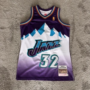 Utah Jazz Karl Malone 32 baskettröja - Retro Utah Jazz baskettröja från Mitchell & Ness, Hardwood Classics-serien. Nummer 32, Karl Malone, säsong 1996-97. Lila och vit med blå och turkosa detaljer, coolt bergsmotiv framtill och broderade loggor. Linnet har tillhört en samling och har därav inte använts. Skriv om det finns frågor!