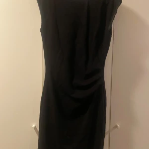 Svart klänning från Tiger of Sweden - En stilren svart klänning från Tiger of Sweden. Modellen är ärmlös och har en smickrande, figurnära passform med drapering på sidan. Perfekt för dig som gillar minimalistisk och klassisk stil.