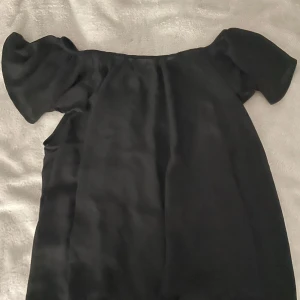 Svart offshoulder topp i satin - Snygg svart offshoulder topp i glansigt satinliknande material. Toppen har korta ärmar och en lös passform som ger en avslappnad men ändå trendig look. Perfekt att styla med jeans eller kjol för en cool vibe.