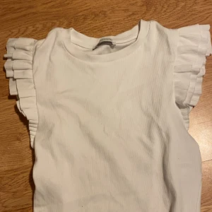 Vit volangtopp från Zara -  vit topp från Zara med ribbad struktur och volangärmar. Toppen är ärmlös med breda, plisserade volanger som ger en extra touch. Mjuk och stretchig känsla. Aldrig använt.