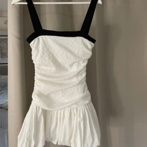 Vit och svart kort klänning Zara S - Supersöt kort klänning från Zara i vit strukturerad bomull med breda svarta axelband och kant upptill. Klänningen har en figurnära passform upptill och en volangig kjol som ger en cool och lekfull vibe. Perfekt för dig som gillar stilrena kontraster och trendig design.