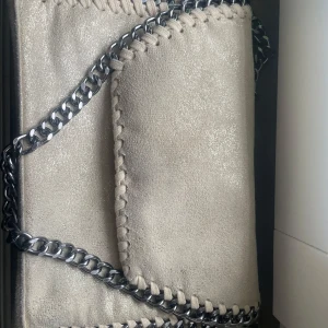 Beige axelväska med kedja - Snygg dope på Stella mcartny beige axelväska i mocka-look med grov silverfärgad kedja som axelrem. Väskan har lock med magnetknäppning och dekorativa stickade detaljer längs kanterna. Insidan har fack med dragkedja för extra förvaring. Perfekt för dig som gillar stilrena accessoarer.