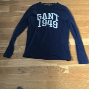 Mörkblå långärmad tröja GANT - Snygg mörkblå långärmad tröja från GANT med vit tryckt text 'GANT 1949' på bröstet. Klassisk rak passform och rund halsringning. Storleken är 158/164 och är oanvänd. Säljes för 200kr.