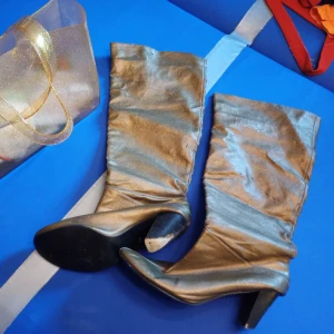 Guldiga stövlar med klack - Snygga guldiga stövlar med hög klack och rund tå. Skorna är i ett glansigt material som ger en riktigt cool look. Perfekta för dig som vill sticka ut och addera lite extra bling till outfiten.