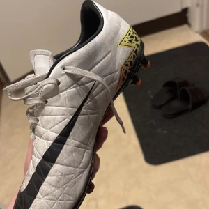 Nike Hypervenom vita fotbollsskor - Säljer ett par Nike Hypervenom fotbollsskor i vitt med stora svarta swoosh-loggor och färgglada detaljer i gult och rosa längs sidorna. Skorna har snörning och mönstrad ovandel i syntetmaterial, samt dobbar för spel på gräs.