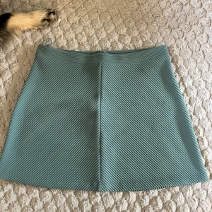 Grön ribbad kortkjol från Lindex - Snygg grön kortkjol från Lindex med ribbat tyg och diskret dragkedja i sidan. Kjolen har en rak passform och diagonala ränder som ger en cool struktur. Perfekt att styla med en enkel topp eller stickad tröja.