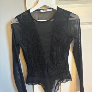 Svart spetsbody från NA-KD - Supersnygg svart body från NA-KD med långärmade meshärmar och djup v-ringning framtill. Framsidan har vackra spetsdetaljer och baksidan är helt i mesh. Den är inte genomskinlig så man behöver inte ha BH under 😇