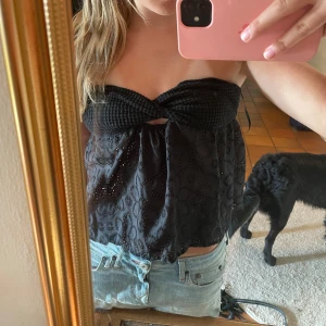 Svart bandeau tubtopp med spetsdetaljer - Supersöt svart bandeau tubtopp med virkad överdel och broderade blommor i spets på tyget. Toppen är croppad och har en liten cut-out framtill. Perfekt för dig som vill ha en unik och trendig look i sommar.